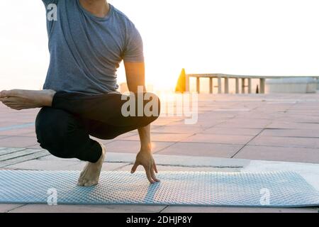 Bilanciamento maschio del raccolto non riconoscibile in posa e facendo Toe Stand yoga a piedi nudi sul tappetino al tramonto in città Foto Stock