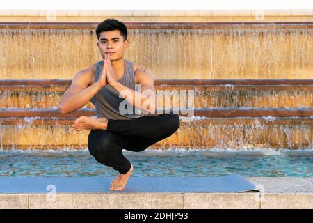 Calzare il maschio asiatico in activewear facendo yoga in Padangusthasana mentre equilibrio sulla gamba sul tappetino e meditazione praticante vicino fontana in città Foto Stock