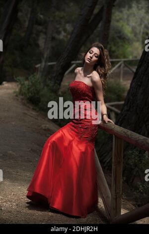 Vista laterale di una donna aggraziata che indossa un lungo abito rosso che pende su recinzione in legno in foresta e godendo della natura con chiuso occhi Foto Stock