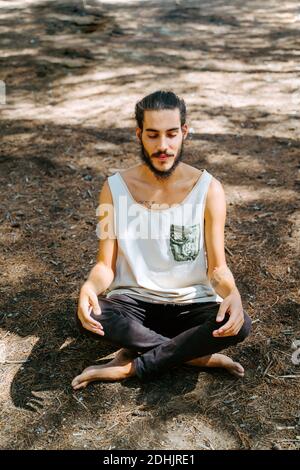 Alto angolo di tranquilla etnia maschile seduta con gambe incrociate In posa di loto e meditando con gli occhi chiusi mentre fa yoga nei boschi Foto Stock