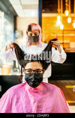 Hairstylist femminile che fa hairdo per il cliente femminile in seduta maschera in salone di bellezza durante l'epidemia di coronavirus Foto Stock