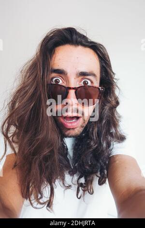 Giovane hipster lungo capelli bearded maschio in occhiali da sole con divertente espressione del volto sbalordita guardando la fotocamera mentre si scattano selfie Foto Stock
