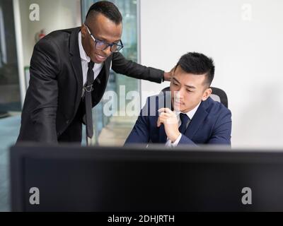 Serio giovane boss asiatico maschio in tuta formale guardando Schermo del portatile con il responsabile afro-americano per spiegare la soluzione del problema durante l'incontro in offi Foto Stock