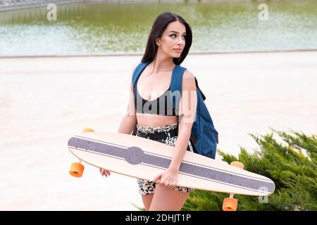Vista laterale di una bella bruna in stile trendy con zaino che trasporta il longboard mentre si è in piedi contro il lungomare urbano in estate giorno Foto Stock