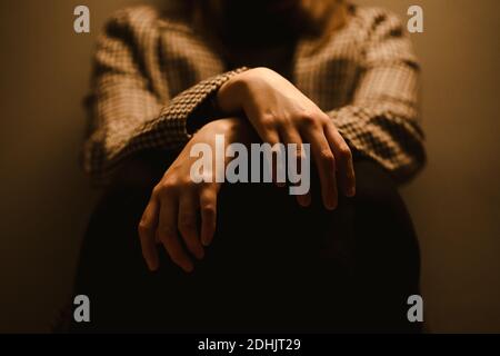 Fuoco selettivo delle mani di raccolto anonimo esaurito depressivo infelice Donna in cappotto a scacchi seduta in studio scuro Foto Stock