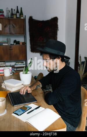 Freelance con vista laterale seduto al tavolo e digitando sul computer portatile mentre si lavora al progetto in un accogliente ufficio domestico Foto Stock