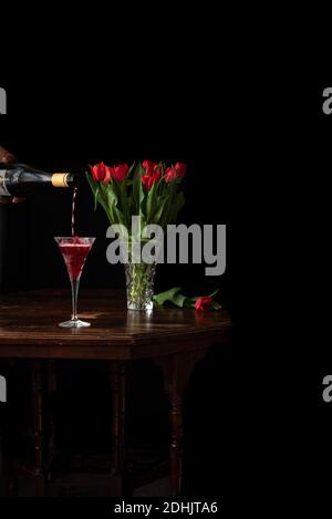 Persona irriconoscibile con bottiglia di vino frizzante in bicchiere sul tavolo con bouquet di tulipani rossi su sfondo nero Foto Stock