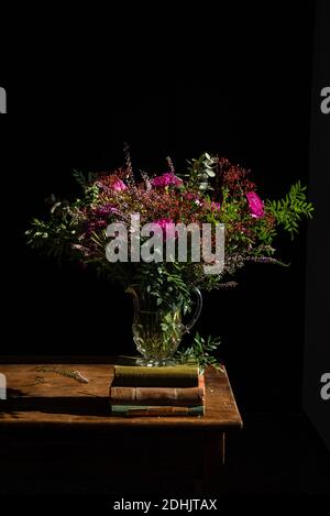 Mazzo di fiori selvatici aromatici assortiti messi in vaso di vetro sopra tavolo rustico in legno con mucchio di vecchi libri su nero sfondo in studio Foto Stock