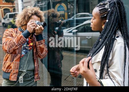 Fotografo maschile creativo con fotocamera di foto di giovani alla moda Modello femminile afroamericano con spirali afro vicino parete di vetro di costruzione urbana Foto Stock