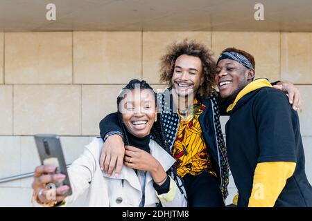 Allegro giovane hipster afroamericano ragazzi abbracciare sorridente ragazza mentre divertirsi insieme sulla scala prendendo selfie con smartphone in città Foto Stock