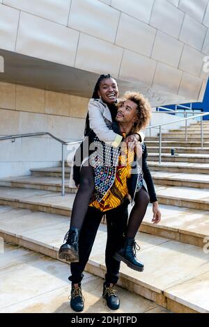 Corpo pieno di allegro giovane ragazzo nero piggybacking ragazza felice mentre ci si diverte insieme sulle scale di pietra vicino alla città moderna edificio Foto Stock