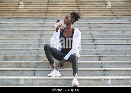 Rilassata afroamericana corridore seduto sulle scale in città e bere acqua fresca durante l'addestramento Foto Stock