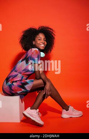 Vista laterale del corpo intero della donna nera con acconciatura afro vestito vivace con sneakers rosa seduto su sfondo rosso Foto Stock