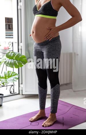Tagliata irriconoscibile concentrata donna incinta in piedi su tappetino e fare esercizi durante l'allenamento domestico Foto Stock