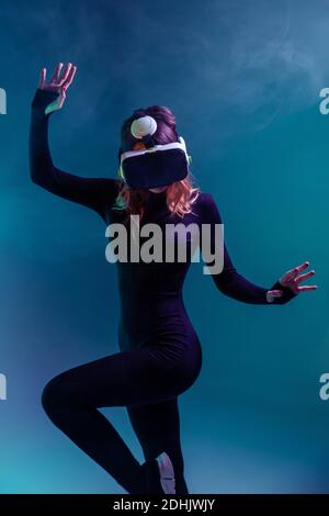 Giovane bionda irriconoscibile Donna che salta a contatto con l'aria e indossa la cuffia per realtà virtuale su sfondo colorato blu neon illuminazione studio Foto Stock