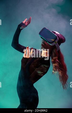 Giovane bionda irriconoscibile Donna in piedi toccare l'aria e indossare la cuffia per realtà virtuale su sfondo colorato blu neon illuminazione studio Foto Stock