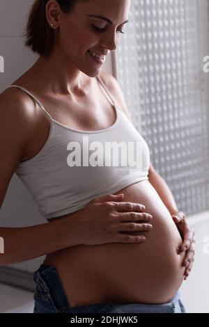 Vista laterale di una bella donna incinta sorridente e delicatamente toccante pancia mentre sei in piedi in camera a casa Foto Stock