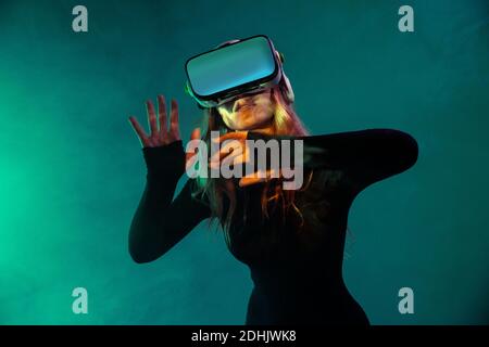 Giovane bionda irriconoscibile Donna in piedi toccare l'aria e indossare la cuffia per realtà virtuale su sfondo colorato blu neon illuminazione studio Foto Stock