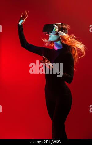 Vista laterale di una giovane donna bionda irriconoscibile che tocca l'aria e indossa una cuffia per realtà virtuale su uno sfondo colorato rosso da studio Foto Stock