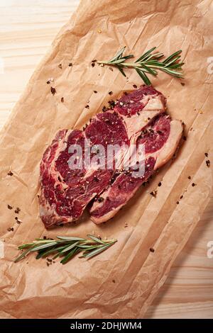 Vista dall'alto della succulenta bistecca di costolette appena sfornata con rosmarino fresco e pepe posto su foglio di carta da forno sul tavolo Foto Stock