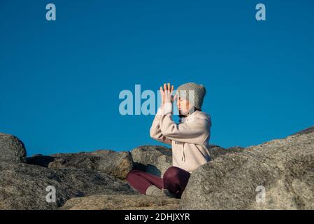 Vista laterale di giovane donna in abiti caldi e lavorato a maglia cappello meditando con le mani di preghiera mentre si siede sulla roccia contro cielo blu Foto Stock