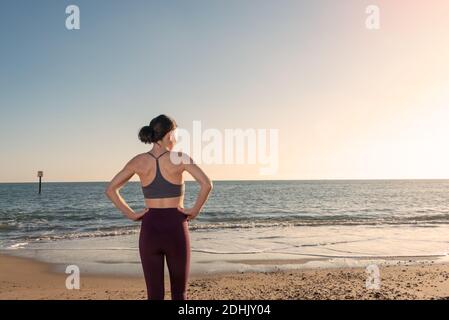 Vista posteriore della femmina sottile in activewear in piedi su sabbia costeggiate vicino all'acqua di mare e godetevi il tempo soleggiato mentre vi rilassate dopo l'allenamento fitness Foto Stock