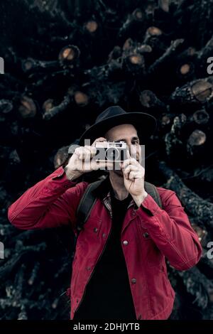 Contenuto uomo adulto in cappello fotografare con foto d'epoca macchina fotografica mentre sta in piedi al mucchio di tronchi di legname Foto Stock