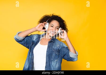 Allegro giovane hipster afroamericano femmina in denim camicia avendo divertimento e ascolto di musica ad alto volume con le cuffie wireless su bright sfondo giallo Foto Stock