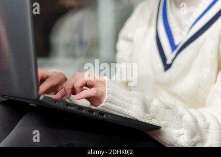 le mani di una ragazza in un maglione caldo stanno battendo testo su un computer portatile a casa vicino alla finestra primo piano Foto Stock