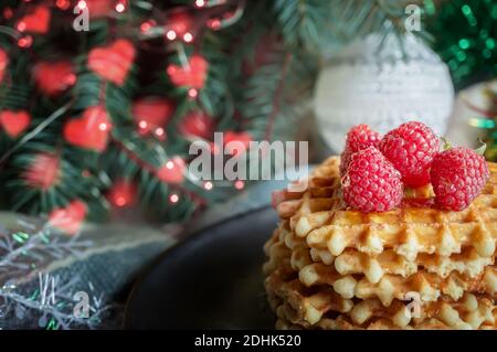 Cialde viennesi con lamponi e miele sul tavolo di Natale con rami di alberi e ghirlande di luci sullo sfondo. I waffle dorati si trovano sopra Foto Stock