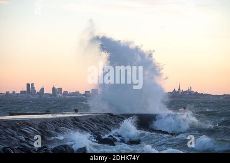 Onde tempestose che si infrangono con un grande spruzzi di acqua frangente sul mare con la silhouette distante della linea della città di Tallinn, che è di colore tramonto Foto Stock