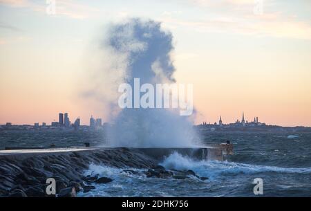 Onde tempestose che si infrangono con un grande spruzzi di acqua frangente sul mare con la silhouette distante della linea della città di Tallinn, che è di colore tramonto Foto Stock