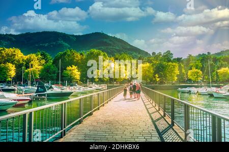 Vista sulla città di Como sul lago di Como il giorno d'estate, yacht e barche, e le persone che camminano verso la riva con alberi e montagne sullo sfondo. Lombar Foto Stock