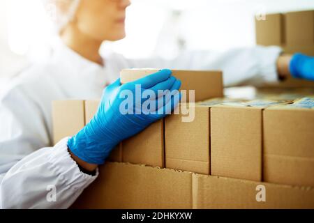 Primo piano di una giovane lavoratrice femminile focalizzata in panni sterili è scatole pieghevoli su pile di scatole nel magazzino di fabbrica. Foto Stock