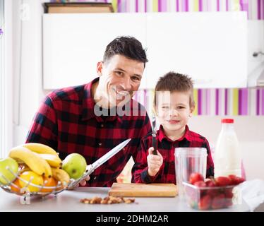 Giovane padre felice e figlio in camicie rosse seduti al banco della cucina sorridendo e tenendo coltelli taglio frutta. Foto Stock
