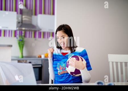 Giovane affascinante ragazza entusiasta in Jersey blu sta guardando una partita di calcio che si acclamano sul suo laptop e tenendo una palla. Foto Stock