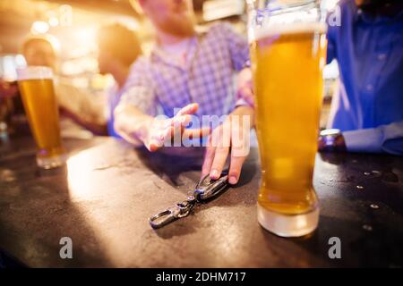 Uomo ubriaco che prende la chiave dell'automobile dal tavolo del pub con una birra alla spina davanti. Primo piano della vista di messa a fuoco di tasti e mani. Foto Stock