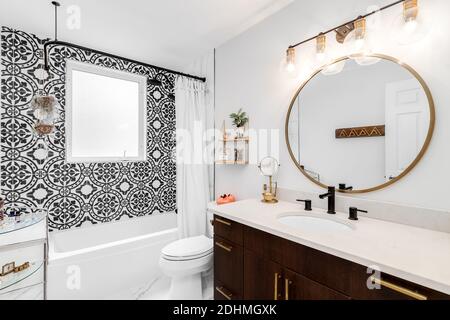A beautifully renovated bathroom with a large gold circular mirror and light fixture, dark wood vanity, and a black and white pattern tiled shower. Stock Photo