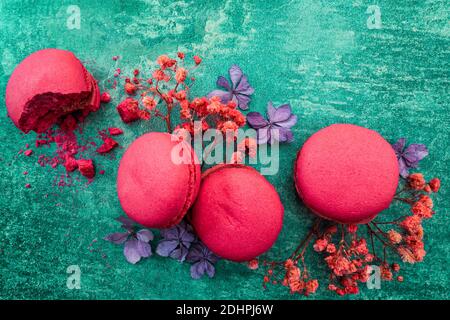 Macaron di fragole su un intenso sfondo verde grunge decorato con fiori. Composizione contrastante disturbata da un biscotto con briciole. Foto Stock