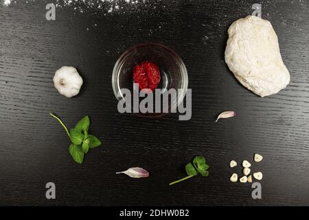 pasta di lievito per la preparazione di pizza, erbe, aglio e pomodoro con spezie giacciono su un tavolo di legno nero Foto Stock