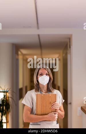 Giovane donna receptionist che tiene la cartella degli appunti con i documenti e con maschera di protezione pandemica in ospedale Foto Stock