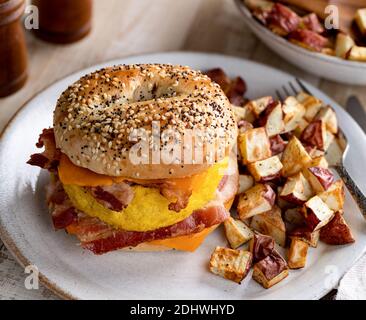 Sandwich con bagel per la colazione a base di uova, pancetta e formaggio cheddar con patate fritte su un piatto Foto Stock