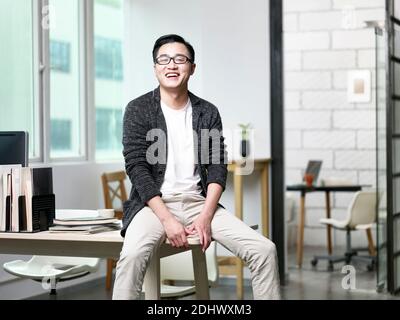 ritratto di giovane uomo d'affari asiatico seduto sulla scrivania in ufficio guardando la macchina fotografica ridendo Foto Stock