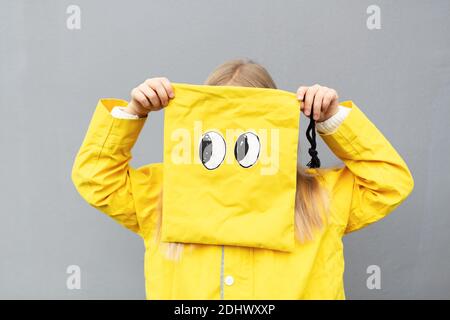 Felice piccola ragazza caucasica 6-7 anni con capelli biondi in impermeabile giallo in piedi all'aperto vicino parete grigia e tenendo borsa divertente. Colori di Foto Stock