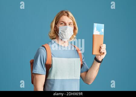 Ritratto del turista in maschera protettiva che tiene in mano i biglietti aerei la sua mano si isolò su sfondo blu Foto Stock