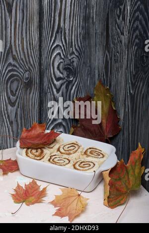 Cottura di cinnabon. L'impasto alla cannella si trova nel piatto da forno. Nelle vicinanze si trovano le foglie d'acero autunnali. Sfondo da assi di pino nero. Foto Stock