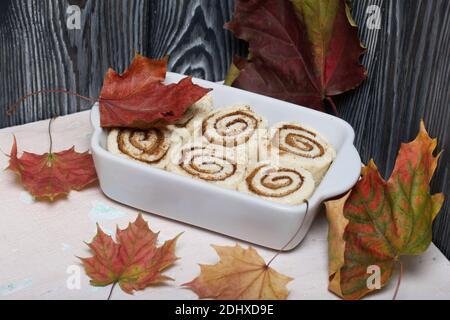 Cottura di cinnabon. L'impasto alla cannella si trova nel piatto da forno. Nelle vicinanze si trovano le foglie d'acero autunnali. Sfondo da assi di pino nero. Foto Stock