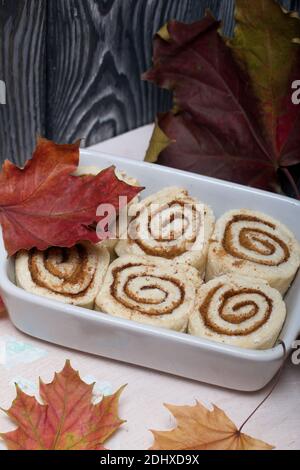 Cottura di cinnabon. L'impasto alla cannella si trova nel piatto da forno. Nelle vicinanze si trovano le foglie d'acero autunnali. Sfondo da assi di pino nero. Foto Stock