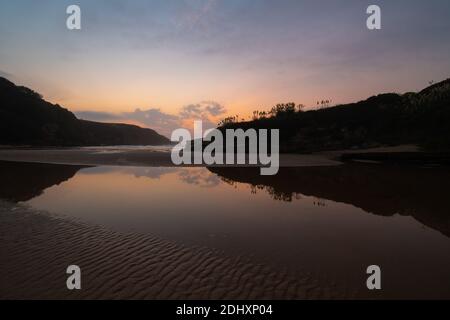 caldo tramonto su una spiaggia con riflessi del cielo in acqua e sagome di vegetazione sulle rocce Foto Stock