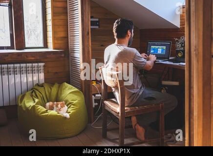 Uomo che lavora al computer a casa, gatto carino sdraiato sulla borsa dei fagioli. Foto Stock